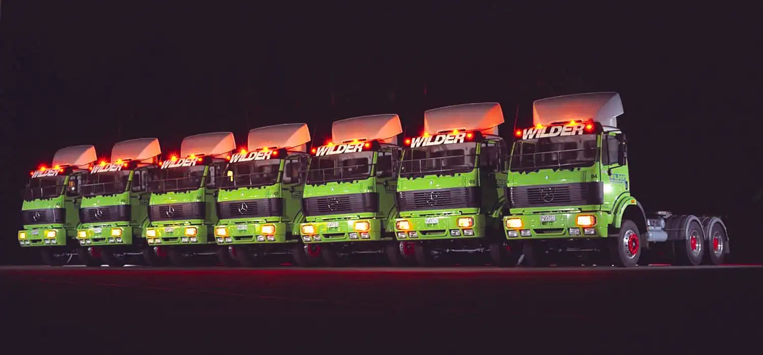 Fleet of green trucks with illuminated cabs parked in a row at night.