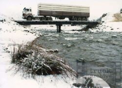 A large truck drives across a bridge over a river surrounded by snow-covered landscape in winter. Heavy truck crossing a bridge over a river in snowy winter conditions.