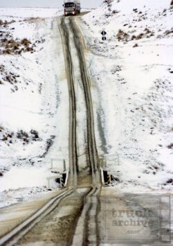 A truck driving on a snowy mountain road with tire tracks and a small bridge crossing in winter. Snow-covered mountain road with truck and tire tracks in winter conditions.