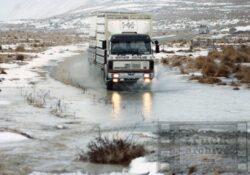 A large truck crossing a flooded rural road in snowy, mountainous terrain. Heavy truck driving through water on a rural road in snowy terrain.