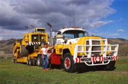 A large yellow mining truck with a loader attached, parked outdoors with a person standing nearby, showcasing heavy equipment. Heavy mining truck and loader on a grassy field under a partly cloudy sky.