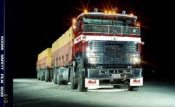 A large Mack truck hauling a long trailer loaded with cargo, illuminated by bright lights during nighttime. Heavy-duty truck at night with bright lights and cargo.