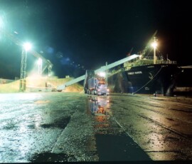 Stock-piles of wood-chips have long been a feature of most of New Zealand's ports - and they got there by truck. Dynes Transport of Tapanui have moved prodigious quantities over six decades, mostly to Port Otago. Cargo ship at port during night with bright lighting and wet ground.